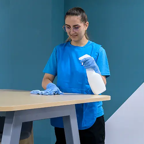 A cleaner in a branded t-shirt wiping a table with a cloth and spray bottle against a teal wall